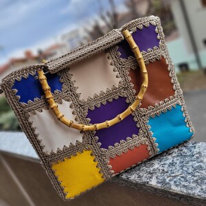 Può includere: Borsa tote fatta a mano con un design patchwork. La borsa presenta quadrati di vari colori, tra cui blu, viola, giallo e marrone, cuciti insieme con un bordo all'uncinetto beige. Ha manici in bambù e una parte superiore all'uncinetto beige.
