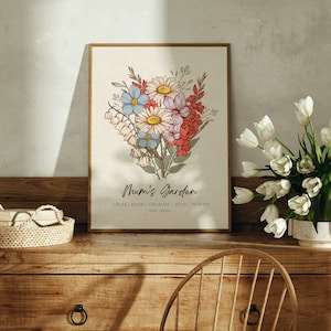 Personalised Mum's Garden birth flower bouquet print in a gold wood frame, styled on a rustic wooden sideboard next to a vase of fresh white tulips, hand-drawn botanical illustration featuring daisies, bluebells, lily of the valley and gladiolus in warm vintage tones, personalised with custom family names and established year, mothers day gift for mum