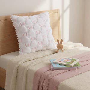 Peut inclure: Un coussin carré blanc avec un motif répété d'oreilles de lapin roses. Le coussin est posé sur un lit avec une couverture crème et un plaid rose. Une figurine de lapin en bois et deux livres pour enfants sont également sur le lit.