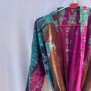 Könnte beinhalten: Ein Batik-Kimono in Türkis-, Braun- und Magentatönen hängt an einem roten Kleiderbügel. Der Kimono hat ein fließendes Design mit langen Ärmeln und einem Bindverschluss. Der Stoff hat ein strukturiertes Aussehen mit einem Muster, das an Aquarellfarben erinnert.
