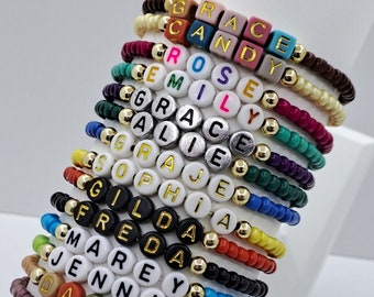 May include: A collection of personalized name bracelets. Each bracelet features colorful beads spelling out a name, with gold-colored accent beads. The bracelets are in various colors, including pink, blue, green, and brown.
