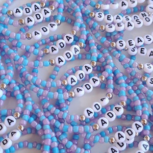 May include: A collection of elastic bracelets made from light blue and lavender seed beads, interspersed with metallic silver spacer beads. Each bracelet features white circular beads with black lettering, spelling out the names ADA or SEASON.