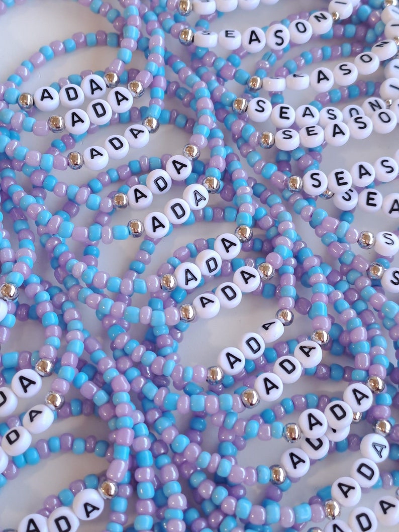 May include: A collection of elastic bracelets made from light blue and lavender seed beads, interspersed with metallic silver spacer beads. Each bracelet features white circular beads with black lettering, spelling out the names ADA or SEASON.
