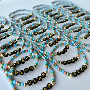May include: A collection of elastic bracelets made with light blue, green, and metallic copper seed beads. Each bracelet features black circular beads with gold lettering that spells out the word BEAR, accented by small gold-toned spacer beads.