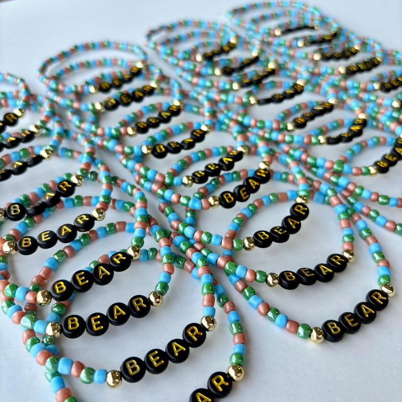May include: A collection of elastic bracelets made with light blue, green, and metallic copper seed beads. Each bracelet features black circular beads with gold lettering that spells out the word BEAR, accented by small gold-toned spacer beads.
