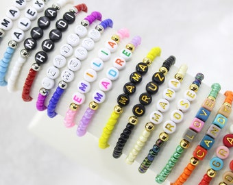 May include: A collection of beaded bracelets with various names spelled out in white and black letter beads. The bracelets feature different colored beads and gold-colored accent beads. Names include "MAMA", "ALEX", "MILA", "HEIDI", "EMILIA", "MADRE", "CRUZ", "ROSE", and "LOVE".