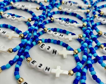 May include: A collection of elastic bracelets made with blue and light blue seed beads, interspersed with small gold-tone metallic beads. Each bracelet features white letter beads spelling E2H and a white cross-shaped bead as a focal point.