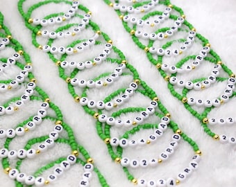May include: Several green beaded bracelets with white letter beads spelling "GRAD 2024" and gold accent beads. The bracelets are arranged in rows, perfect for a graduation celebration.