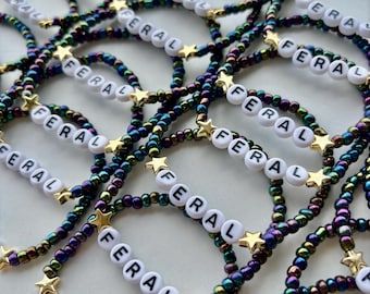 May include: A collection of beaded bracelets with the word "FERAL" spelled out in white letter beads. The bracelets feature iridescent beads in shades of blue, green, and purple, accented with gold star charms.