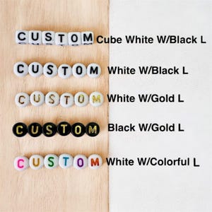 May include: Five rows of alphabet beads spelling CUSTOM. Row one: white cube beads with black letters. Row two: white round beads with black letters. Row three: white round beads with gold letters. Row four: black round beads with gold letters. Row five: white round beads with colorful letters.