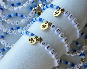 Könnte beinhalten: Eine Sammlung von Perlenarmbändern mit weißen Buchstabenperlen, die BEST DAY EVER buchstabieren, durchsetzt mit blauen und weißen Saatperlen. Jedes Armband enthält einen kleinen goldfarbenen Sternanhänger und einen runden goldfarbenen Anhänger mit Logo.