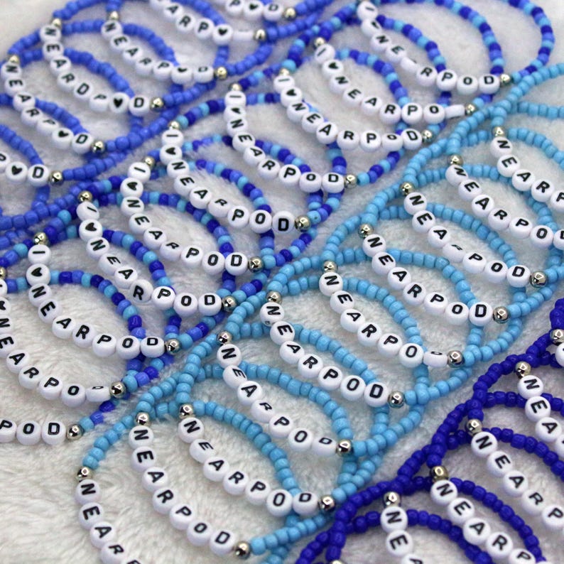 May include: A collection of elastic bracelets made from small blue and light blue seed beads. Each bracelet features white spherical beads with black letters spelling out the word NEARPOD, interspersed with small metallic silver spacer beads.