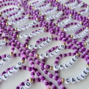 May include: A collection of elastic bracelets made from small purple and light purple beads, interspersed with metallic gold beads. Each bracelet features white letter beads spelling the word CALLED in black, arranged in a repeating pattern.