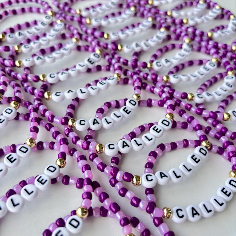 May include: A collection of elastic bracelets made from small purple and light purple beads, interspersed with metallic gold beads. Each bracelet features white letter beads spelling the word CALLED in black, arranged in a repeating pattern.