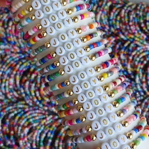 May include: A stack of colorful beaded bracelets featuring white circular beads with the word JOY printed in gold. Each bracelet alternates between small multi-colored beads and metallic gold spherical beads, creating a vibrant pattern.