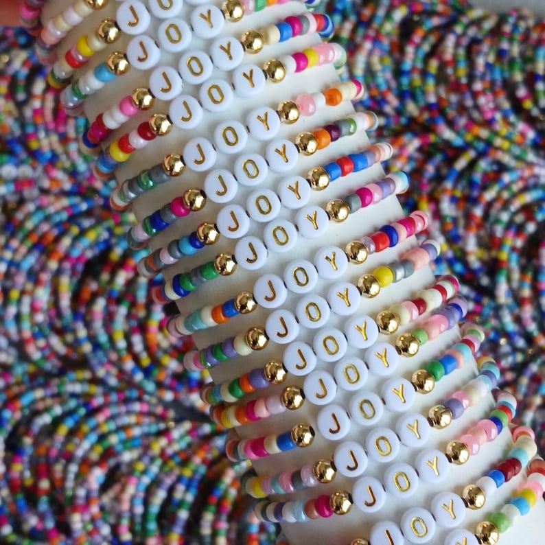 May include: A stack of colorful beaded bracelets featuring white circular beads with the word JOY printed in gold. Each bracelet alternates between small multi-colored beads and metallic gold spherical beads, creating a vibrant pattern.