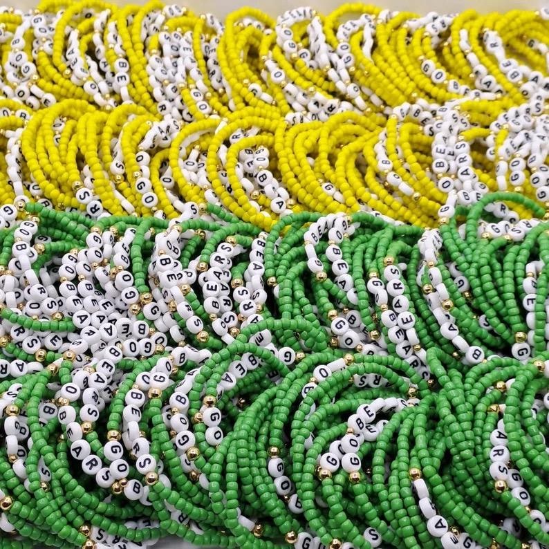 May include: A collection of elastic bracelets made from small yellow or green beads, interspersed with white letter beads spelling GO BEARS and small metallic gold spacer beads. The bracelets are arranged in a dense, layered group.