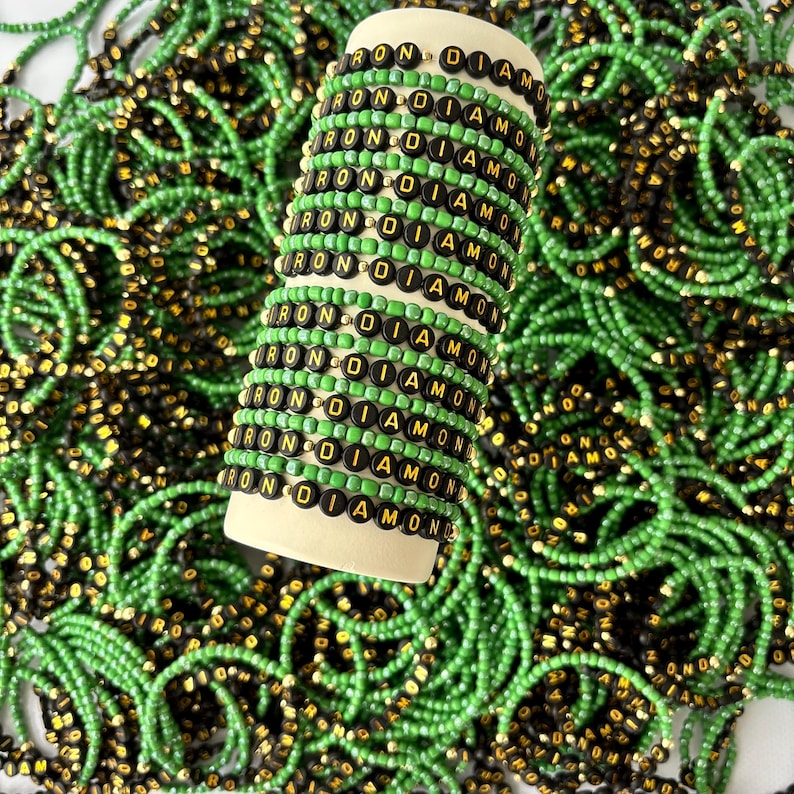 May include: A collection of elastic bracelets featuring alternating rows of bright green seed beads and black circular beads with the gold-colored text IRON DIAMOND printed on them, stacked on a cylindrical display stand.