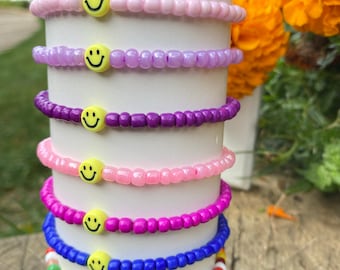 May include: A stack of eight beaded bracelets with smiley face charms. The bracelets are in various colors, including red, pink, purple, blue, and black.