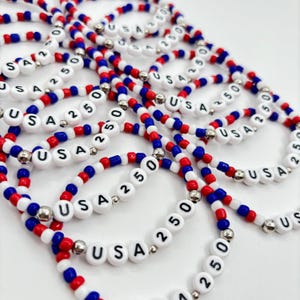May include: A collection of elastic bracelets made from red, white, and blue beads. Each bracelet features white letter beads spelling USA and number beads reading 250, interspersed with small metallic silver spacer beads throughout the design.