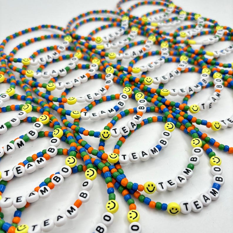 May include: A collection of elastic bracelets made from small, multicolored beads in blue, green, and orange. Each bracelet features white letter beads spelling out the text TEAM BO, interspersed with yellow smiley face beads throughout.