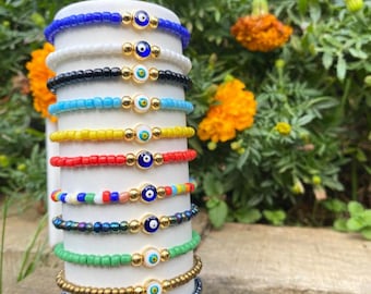 May include: A stack of 12 colorful beaded bracelets with gold accents and blue evil eye charms. The bracelets are stacked on a white ceramic stand.