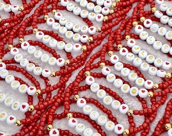 May include: Red beaded bracelets with white lettered beads spelling "JLGO" and heart-shaped beads. The bracelets have gold-colored accent beads. The bracelets are displayed in a close-up shot.