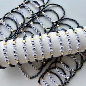 May include: A collection of beaded bracelets featuring iridescent dark-toned seed beads, small gold-colored star charms, and white round letter beads spelling out the word FERAL. The bracelets are arranged in a pile and draped over a white cylinder.