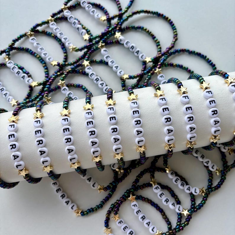 May include: A collection of beaded bracelets featuring iridescent dark-toned seed beads, small gold-colored star charms, and white round letter beads spelling out the word FERAL. The bracelets are arranged in a pile and draped over a white cylinder.