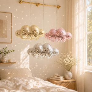 May include: Three cloud-shaped disco ball pendant lights in gold, pink, and silver hang from a gold rod. The lights are suspended above a bed with a cream-colored comforter. The room has a warm, inviting atmosphere.