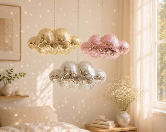 May include: Three cloud-shaped disco ball pendant lights in gold, pink, and silver hang from a gold rod. The lights are suspended above a bed with a cream-colored comforter. The room has a warm, inviting atmosphere.