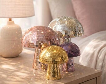May include: A collection of decorative mushroom-shaped lamps with mirrored mosaic surfaces. The lamps are in various colors, including gold, rose gold, silver, and purple. They are arranged on a wooden surface, with a lamp and bedding in the background.