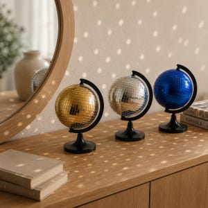 Peut inclure: Trois boules à facettes en forme de globe montées sur des supports noirs, disposées en rangée. Les globes sont recouverts de petites tuiles carrées réfléchissantes dorées, argentées et bleues, créant des reflets sur le mur et la surface.