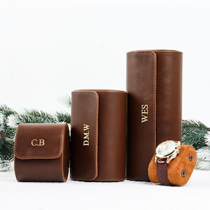 Könnte beinhalten: Drei braune Leder-Uhrenetuis in verschiedenen Größen, jeweils mit goldfarbenen Buchstaben. Das kleinste Etui ist quadratisch, das mittlere zylindrisch und das größte ebenfalls zylindrisch. Eine Uhr mit braunem Armband liegt auf einem braunen Wildleder-Kissen.