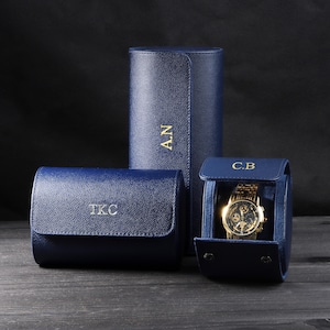 Puede incluir: Tres estuches de reloj azul marino con iniciales doradas. Un estuche cilíndrico y dos estuches rectangulares. Un estuche rectangular está abierto, mostrando un reloj dorado con una correa dorada. Las iniciales "A.N", "T.K.C" y "C.B" son visibles.