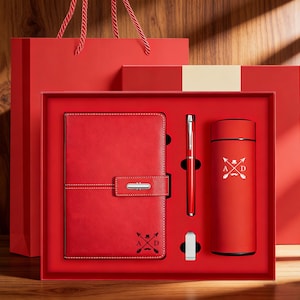 May include: A red gift set containing a notebook, pen, tumbler, and a small metal clip, presented in a red box and bag. The notebook has a silver clasp and white stitching. The tumbler features a white graphic. A stylish gift set for any occasion.
