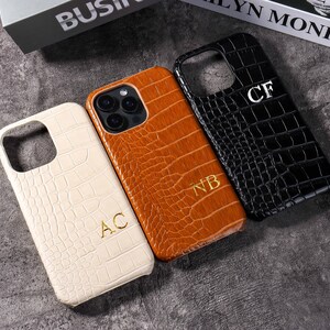 Könnte beinhalten: Drei personalisierte Handyhüllen in Creme, Braun und Schwarz, jeweils mit Krokodilhautstruktur. Die cremefarbene Hülle hat die Initialen "AC", die braune "NB" und die schwarze "CF".