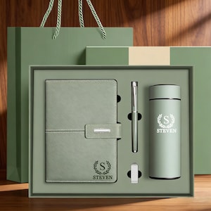 Puede incluir: Un juego de regalo verde salvia que incluye un cuaderno, un bolígrafo, un vaso y una unidad USB. El cuaderno tiene un cierre de solapa y el vaso está grabado con el nombre "STEVEN". El juego se presenta en una caja de regalo con una bolsa a juego.