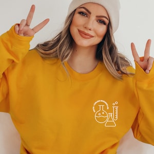 May include: Mustard yellow sweatshirt with a white graphic of laboratory glassware and the text "SMITH + CARLSEN RESEARCH LAB". The sweatshirt is a crew neck style.