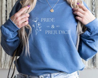 Puede incluir: Una sudadera azul de cuello redondo con una ilustración floral de líneas blancas en el lado izquierdo y el texto blanco PRIDE est. & 1813 PREJUDICE impreso en el frente. La prenda tiene mangas largas y cuello, puños y dobladillo acanalados.