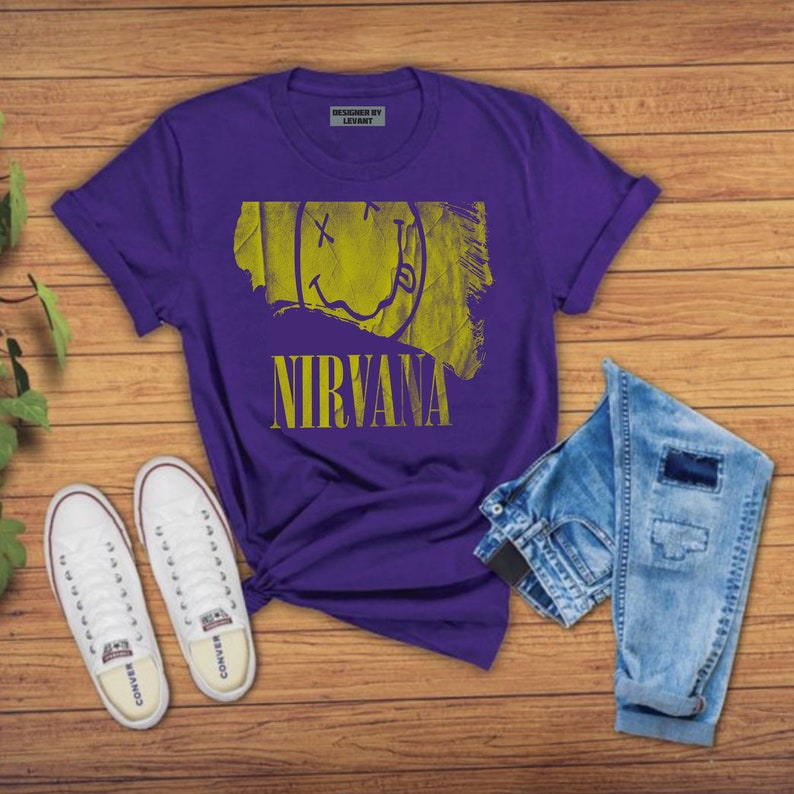 Pode incluir: Camiseta roxa com um gr&aacute;fico de rosto sorridente amarelo e a palavra "NIRVANA" impressa na frente. A camiseta est&aacute; colocada sobre uma superf&iacute;cie de madeira com um par de t&ecirc;nis brancos e uma cal&ccedil;a jeans azul.