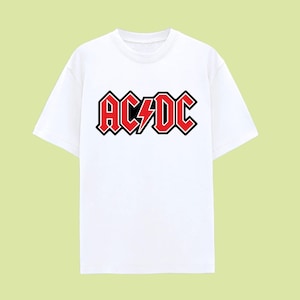 May include: White t-shirt featuring the band name "AC/DC" in red, outlined in black, with a lightning bolt graphic. The text is in a bold, stylized font. The shirt has short sleeves and a crew neck.