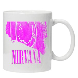 Pode incluir: Caneca de cer&acirc;mica branca com um design grunge rosa que apresenta um rosto sorridente com um X para os olhos e a palavra "NIRVANA" abaixo.