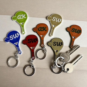 May include: A selection of tennis racket-shaped keyrings in various colours, including green, blue, red, orange, and gold. Each keyring features a tennis ball and initials. Some have silver key rings attached. Keys are also present.