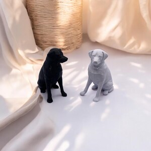 Puede incluir: Dos figuras de perro, una negra y otra gris, est&aacute;n sobre una superficie blanca. Los perros est&aacute;n sentados, con las orejas erguidas. Una cesta tejida y tela drapeada se encuentran en el fondo.