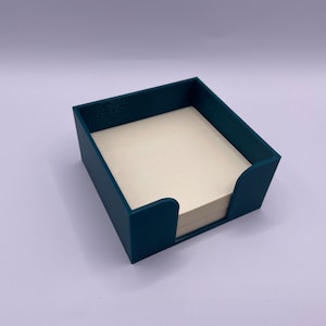Peut inclure: Un organisateur de bureau bleu sarcelle pour des notes adhésives. L'organisateur est en plastique et a une forme carrée. Les notes adhésives sont blanches et sont visibles à l'intérieur de l'organisateur.
