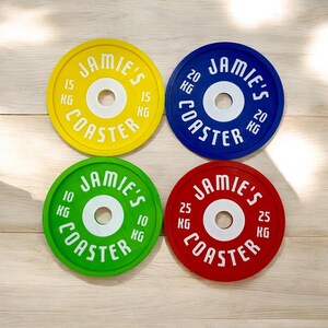 Puede incluir: Cuatro posavasos circulares dise&ntilde;ados como discos de pesas en amarillo, azul, verde y rojo. Cada posavasos tiene un orificio central blanco y el texto blanco JAMIE'S COASTER con marcas de peso de 15 KG, 20 KG, 10 KG y 25 KG.
