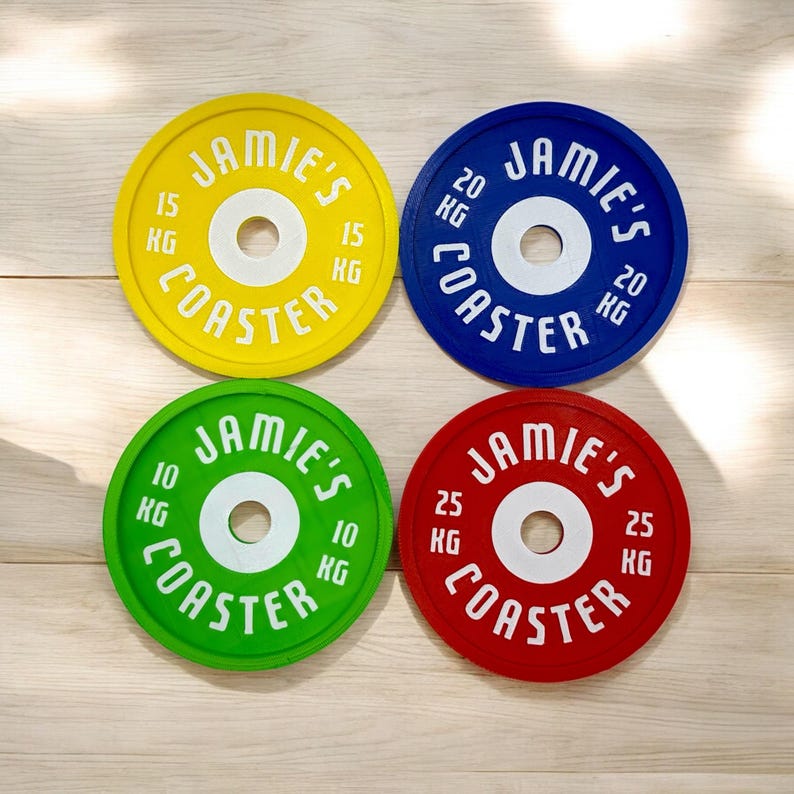 Puede incluir: Cuatro posavasos circulares dise&ntilde;ados como discos de pesas en amarillo, azul, verde y rojo. Cada posavasos tiene un orificio central blanco y el texto blanco JAMIE'S COASTER con marcas de peso de 15 KG, 20 KG, 10 KG y 25 KG.