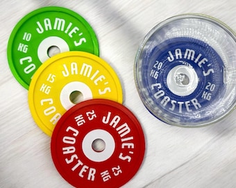May include: A collection of circular coasters designed to resemble weight plates. The coasters are in shades of green, yellow, red, and blue. Each coaster features the text "JAMIE'S COASTER" and weight measurements in kilograms.