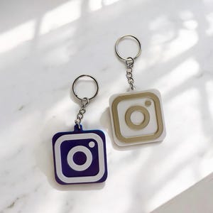 May include: Two keychains with Instagram logo charms. One charm is white with a gold outline and the other is purple with a white outline.
